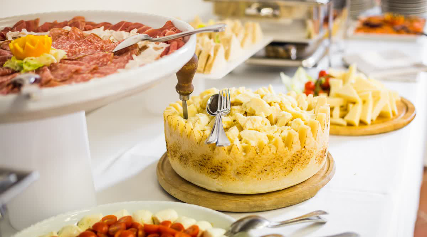 Una tavola imbandita con una varietà di cibi. In primo piano a sinistra, un vassoio bianco rialzato contiene affettati misti disposti artisticamente. Al centro, un'ampia forma di formaggio scavata è riempita di cubetti di formaggio, con posate inserite. Più a destra, un tagliere di legno ospita bastoncini di formaggio. Si intravedono anche altri piatti con mozzarella e pomodorini.